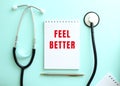 On a blue background, a stethoscope and a white notepad with the red words FEEL BETTER. Royalty Free Stock Photo