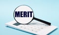 On a blue background, a white calculator and a magnifying glass with the text MERIT. Business concept Royalty Free Stock Photo