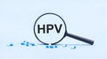 On a blue background, white and blue capsules with pills and a black magnifying glass with the text HPV. Medical concept