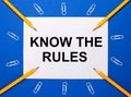 On a blue background, white paper clips, yellow pencils and a white sheet of paper with the text KNOW THE RULES Royalty Free Stock Photo