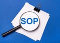 On a blue background, white sheets under a black paper clip and on them a magnifying glass with the text SOP Standard Operating Royalty Free Stock Photo