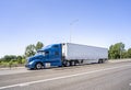 Blue big rig classic semi truck with high cab transporting load in refrigerator semi trailer running on the highway road entrance Royalty Free Stock Photo