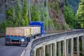 Blue big rig classic semi truck transporting lumber wood on flat bed semi trailer driving on the bridge on the mountainside in Royalty Free Stock Photo