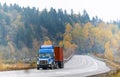 Blue big rig semi truck with flat bed semi trailer transporting container running on winding autumn road with raining weather