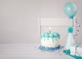 Blue Birthday cake, presents, hats and balloons over light grey background.