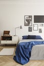 Blue blanket on bed in bright bedroom interior with lamp on cabinet near posters on white wall. Real photo Royalty Free Stock Photo