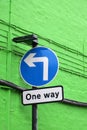 Blue circular road sign with a left turn arrow and notice saying one way against a green brick wall Royalty Free Stock Photo