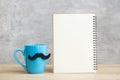 Blue coffee cup or tea mug with Black mustache decor and empty paper notepad or calendar on table. Blank copy space for text. Blue Royalty Free Stock Photo