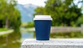 Blue coffee cup with white lid outdoors by a pond with trees and mountains in background Royalty Free Stock Photo