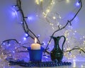 Blue composition with candle and miniature mug, blue pearl ornaments in the foreground, white and blue light chain background, Royalty Free Stock Photo