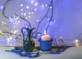 Blue composition with candle and miniature mug, blue pearl ornaments in the foreground, white and blue light chain background, Royalty Free Stock Photo