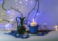 Blue composition with candle and miniature mug, blue pearl ornaments in the foreground, white and blue light chain background, Royalty Free Stock Photo