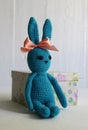 A blue crocheted banny rabbit sits near a gift box on a white background