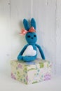 A blue crocheted banny rabbit sits on a gift box on a white background