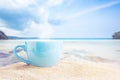 Blue cup of coffee on white sand beach over blue sky and sea on day noon light background.