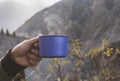 Blue cup with hot tea or coffee with steam over it in a hand. Mountain view in the background.