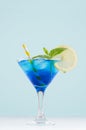 Blue curacao cocktail with ice cubes, lemon slice, yellow straw, green mint in luxury martini glass on white wood board and green. Royalty Free Stock Photo