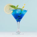 Blue curacao cocktail with ice cubes, lemon slice, yellow straw, green mint in luxury martini glass on white wood board and pastel Royalty Free Stock Photo