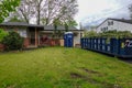 Blue dumpster filled with tree evergreen branches and a blue outhouse in the driveway of a house without siding