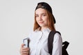 Blue eyed delighted schoolgirl holds book, carries backpack, wears black cap, going to library, reads and cramms material for exam