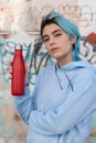 Blue haired Teenage girl in blue hoodie staying near graffiti wall with red water bottle Royalty Free Stock Photo