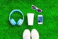 Blue headphones, white smartphone, cup of fruit juice with sneakers shoes, sunglasses on a green grass textured background