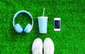 Blue headphones, white smartphone, cup of fruit juice with sports sneakers shoes on a green grass textured background