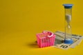 Blue hourglass and dollars with shopping basket at yellow background