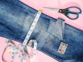 Blue jeans with large hole folded in half, sewing pins in a box, white tailor tape with centimeters and inches and scissors.