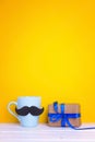 Blue mug with a mustache and gift box on yellow background. Copy Royalty Free Stock Photo