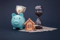 Blue piggy bank with wooden model of house, hourglass and money banknotes on blue background. Savings money for buy