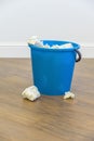 A blue plastic bucket filled with used tissues sits on a wooden floor