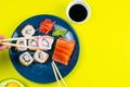 Blue plate with sushi, filled with rolls of different kinds, next to stand sticks for sushi, red marinaginger and soy sauce