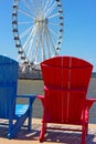 Blue and red chairs on a pier with Ferris wheel on background.