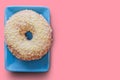 On a blue saucer is a sweet, fresh doughnut with a white glaze. The view from the top. Pale pink background. Free space for