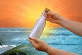 Blue sea, glass bottle with message in female hands, fast waves to shore, beautiful tropical sunset reflected in water, leaves Royalty Free Stock Photo