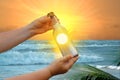 blue sea, glass bottle with message in female hands, fast waves to shore, beautiful tropical sunset reflected in water, leaves Royalty Free Stock Photo
