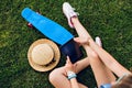 Blue skateboard, hat, legs and tablet in hands of girl sitting on grass in park