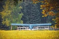 Blue small airplane parked in field near white wooden fence with blurred background of evergreen forest
