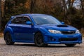 Blue subaru wrx car parked in a dirty parking lot with fall foliage in the background