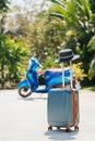 Blue SUITCASE with a hat on the handle, stands on the street, against a background of tropical palm trees and a scooter.
