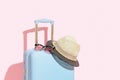 Blue suitcase with sunglasses and hat on a pastel pink background with long deep shadows. Travel concept