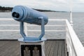 Blue telescope mounted on a stand on wooden ocean pier with white railing. Cloudy summer day on the pier of Jurata