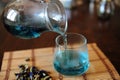 Blue Thai tea anchan is poured from jug into glass. Placer of dried flowers of clitoria tea