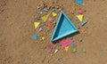 blue triangular object lies amidst scattering multicolored triangular pieces on sandy surface.
