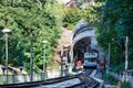 A blue and white cable funicular picks up passengers at the lower station, surrounded by a summer green park