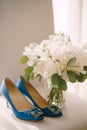 Blue women`s shoes with a buckle toe on a white surface with a bouquet of peonies in a vase.