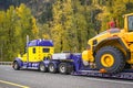 Blue and yellow powerful big rig semi truck transporting oversize load on heavy duty step deck semi trailer driving on the autumn Royalty Free Stock Photo