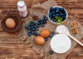 Blueberries with leaf in the white bowl, flour in the bowl, wooden spoon, yogurt, eggs on brown wooden background