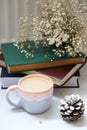 a bluish gray and pink cup of coffee and white colored pine cone and bunch of tiny flowers on books and a curtain